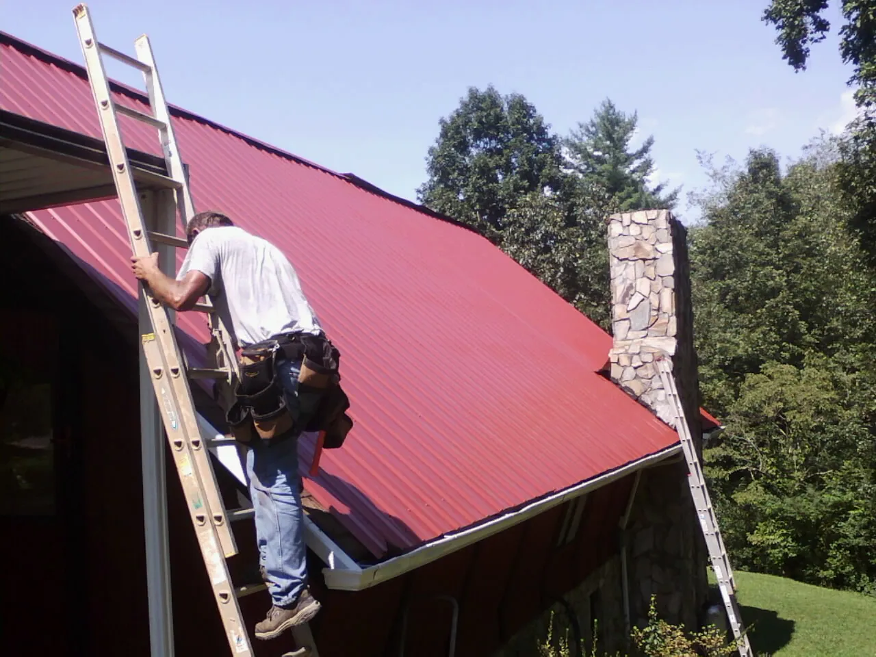 Roofer on a ladder beside a red metal roof and stone chimney — Soffit & Fascia Repair by Roofing Brothers in Villisca